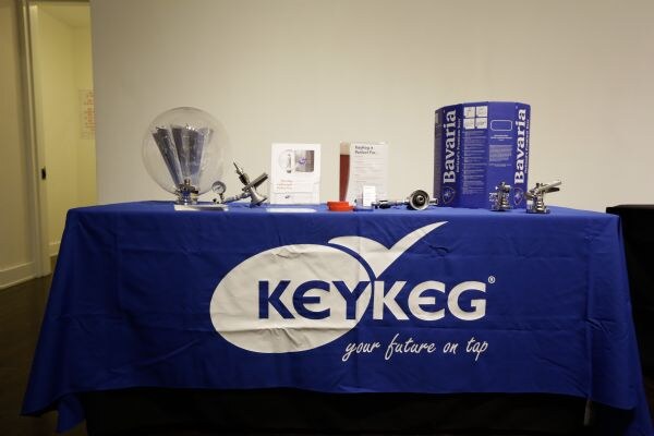 A table with a blue KeyKeg tablecloth holds a glass dome, printed cards, metal couplers and a blue Bavaria keg carton, in front of a plain white wall.