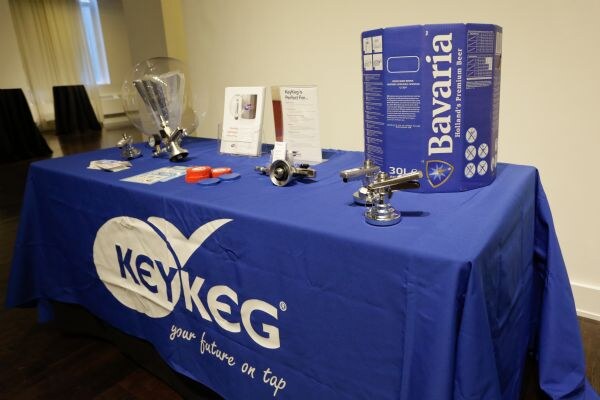 Angled view of a table with a blue KeyKeg tablecloth holding a Bavaria 30-liter keg carton, metal couplers, a glass dome and printed cards.