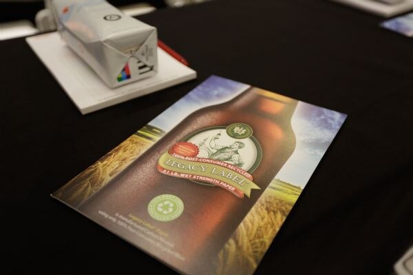 A Legacy Label brochure showing a beer bottle over a wheat field, labeled 100% post-consumer recycled, lies on a black table beside a notepad and a small folded paper carton.