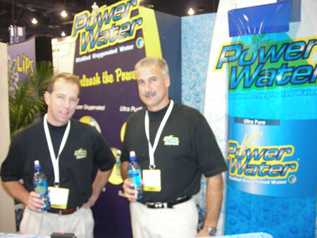 Two men in black shirts with badges hold bottles of Power Water in front of Power Water banners, including a tall bottle graphic on the right.