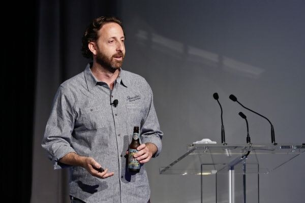 A bearded man in a gray button-up shirt with a Shmaltz Brewing logo and a lapel microphone holds a beer bottle and a clicker while speaking on stage near a clear podium.