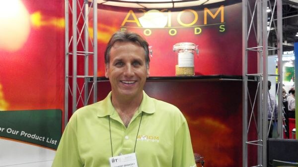 A smiling man in a lime green polo shirt with an Axiom logo and an IFT badge stands in front of the Axiom Foods booth, which has a jar of grain on a shelf and truss framing.