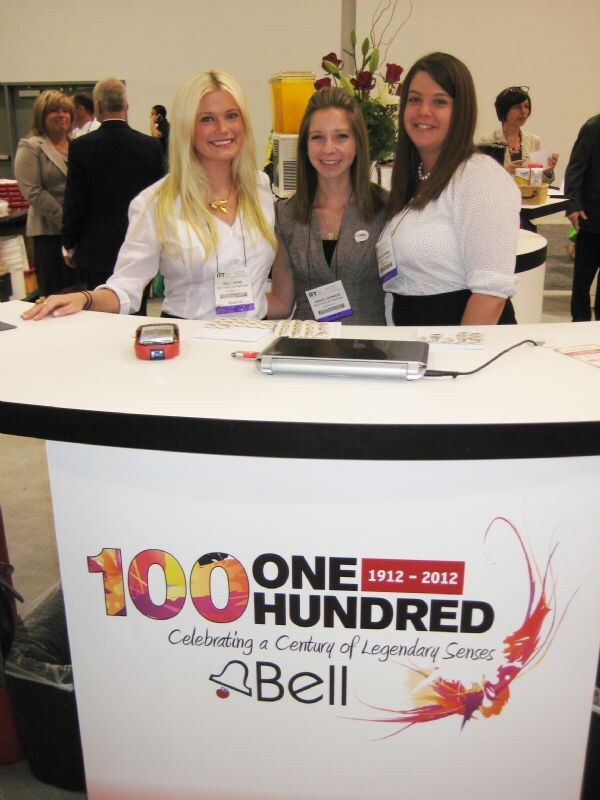 Three smiling women with IFT badges stand behind a white Bell booth counter with a laptop on it. The counter reads 100 One Hundred, Celebrating a Century of Legendary Senses, 1912 to 2012.