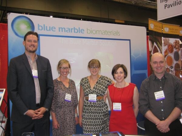 Five people with name badges stand in front of a Blue Marble Biomaterials display at a trade show booth: a bearded man in a suit, three women, and a bald man in a striped shirt.