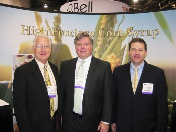 Three men in dark suits with IFT badges stand in front of a display reading High Fructose Corn Syrup, with corn imagery and a Bell sign overhead.