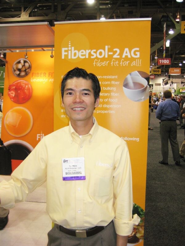 Tu Tran, in a yellow button-up shirt with an IFT badge, smiles in front of an orange Fibersol-2 AG banner reading fiber fit for all.