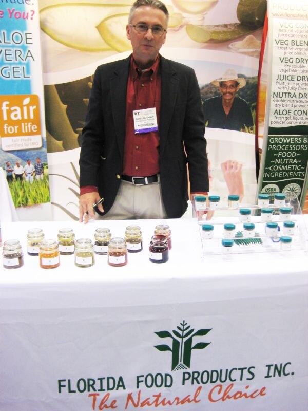 A man in a dark blazer and red shirt stands behind a table with small jars of ingredient samples and a Florida Food Products Inc. tablecloth, with aloe vera banners behind him.