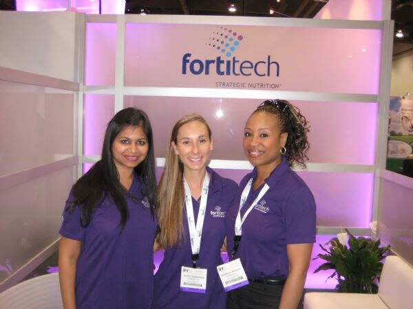 Three smiling women in purple Fortitech polo shirts stand in front of a lit pink Fortitech Strategic Nutrition sign. Two wear IFT badges.