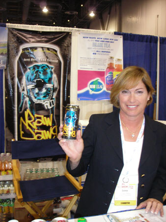 A smiling woman with short light brown hair and a dark blazer holds up a can of Raw Dawg in a booth with a Raw Dawg banner and a New Leaf Blue Tea poster behind her.