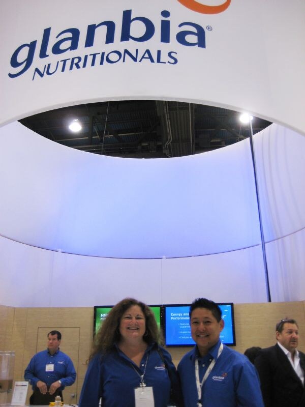A woman and a man in blue Glanbia shirts smile under a large Glanbia Nutritionals sign. Two other men stand in the booth behind them.