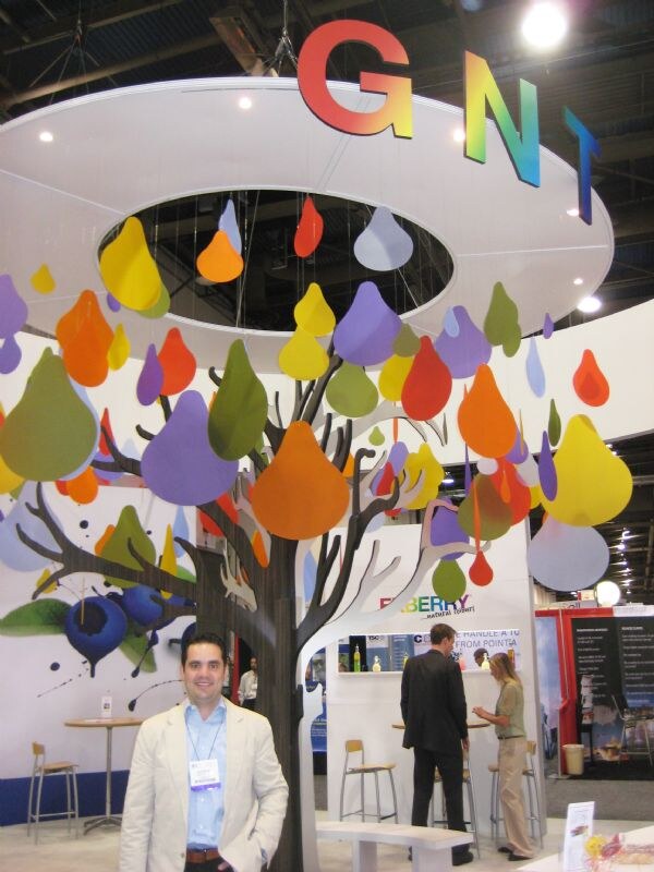 A smiling man in a light jacket with a badge stands before the GNT booth, which has a tree sculpture with colorful teardrop shapes. Two people talk at a counter behind him.