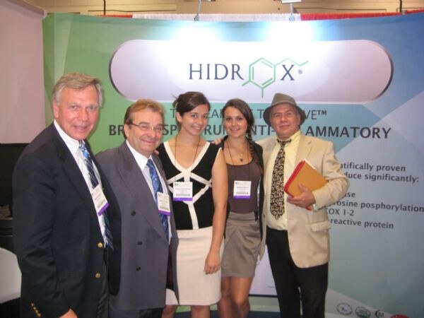 Five people, three men and two women, pose in front of a Hidrox banner. Most wear badges, and one man wears a gray hat and holds an orange folder.