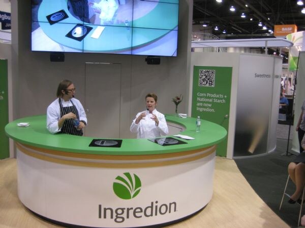 Two people in chef coats stand behind a green and white Ingredion demonstration counter, one speaking. A large screen is above, and a sign says Corn Products + National Starch are now Ingredion.
