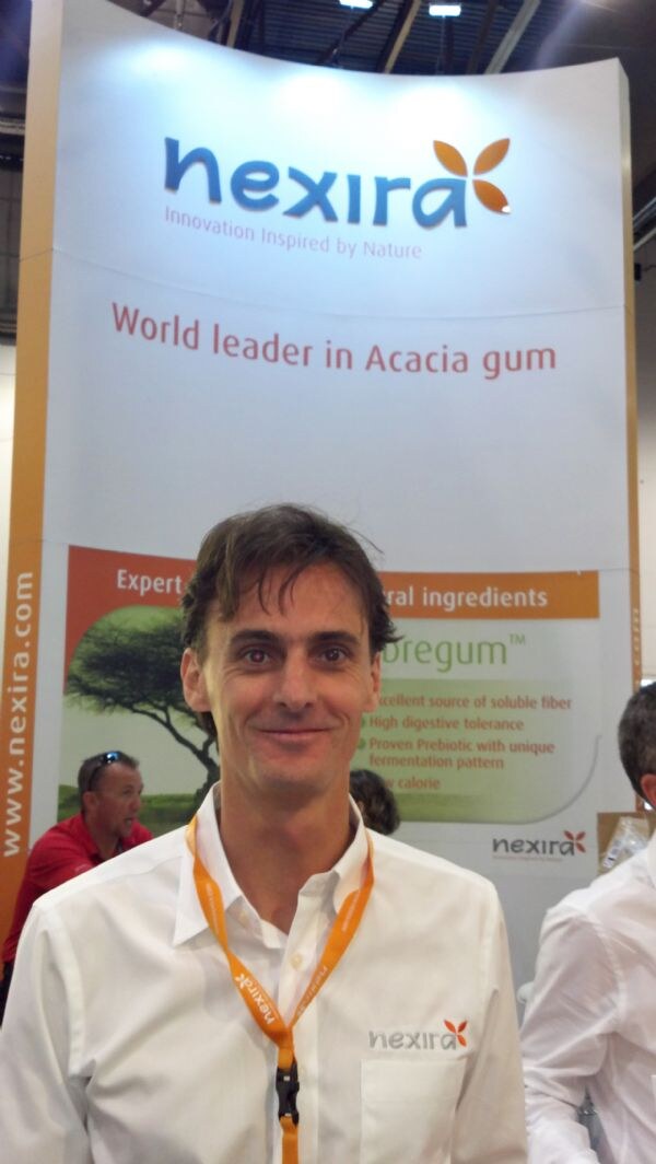 A smiling man in a white Nexira shirt and orange lanyard stands before a Nexira banner reading World leader in Acacia gum.