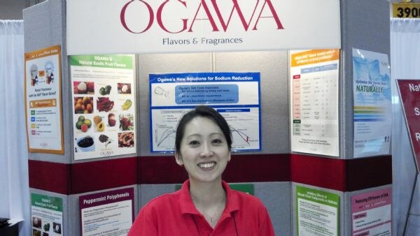 A smiling woman in a red shirt stands in front of the Ogawa Flavors & Fragrances booth, with posters about fruit flavors and sodium reduction behind her.