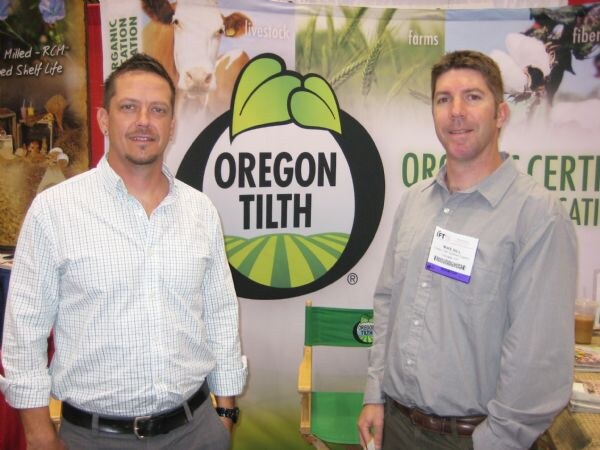 Two men stand smiling in front of an Oregon Tilth banner with its green logo. The man on the right wears an IFT badge.
