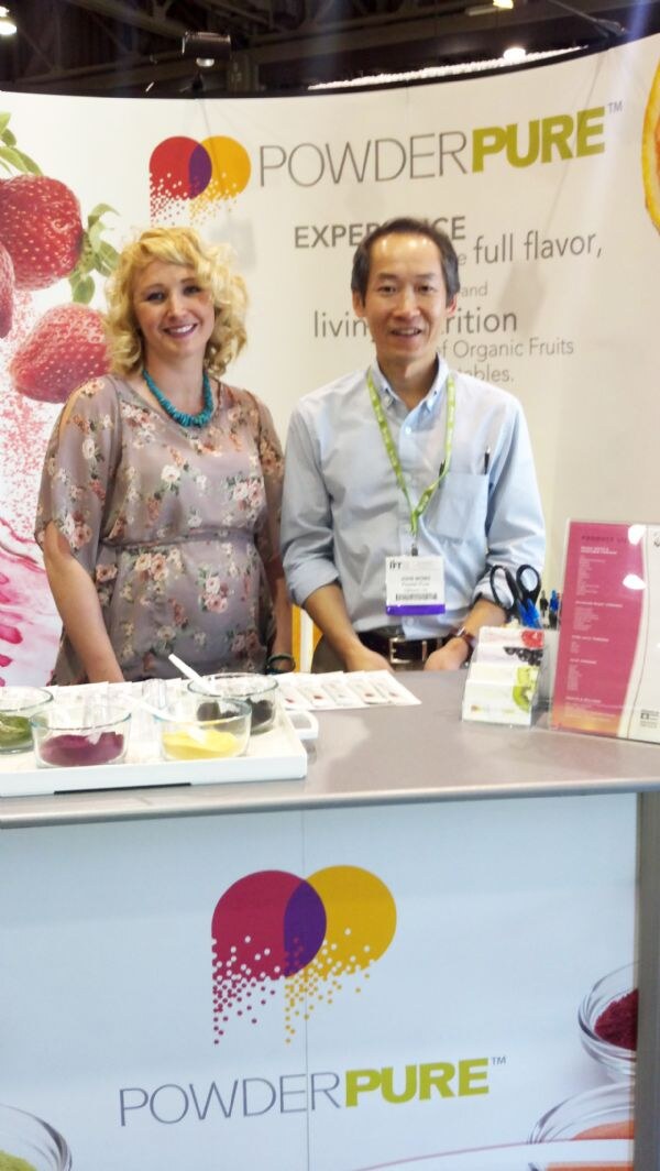 A blonde woman and a man with a green lanyard stand behind a PowderPure booth counter with small dishes of colorful fruit powders and a banner showing strawberries.