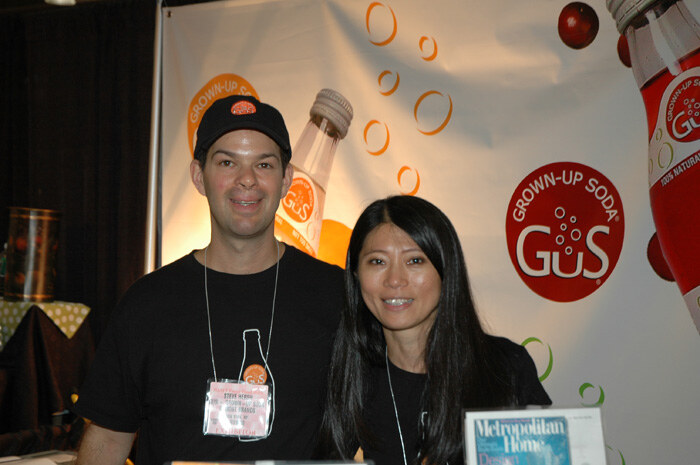 A man in a black cap and a woman with long dark hair stand smiling in front of a GuS Grown-Up Soda banner with bottle graphics. A Metropolitan Home magazine sits in the foreground.