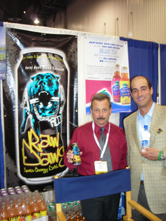 Two men stand beside a director's chair at a booth. One in a red shirt holds a Raw Dawg can; the other in a tan jacket holds a small bottle. A Raw Dawg banner and New Leaf poster are behind them.