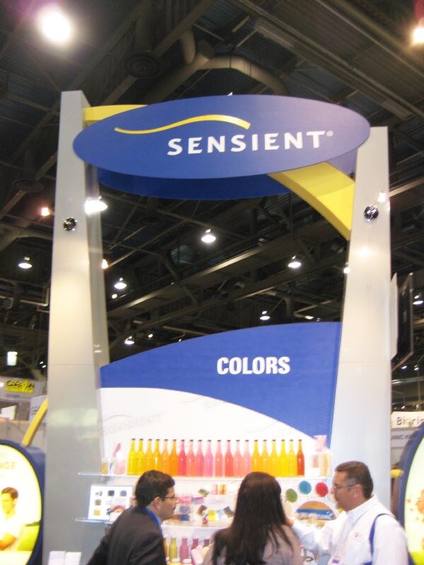 A tall blue and gray Sensient booth with a Colors sign above a shelf of bottles in yellow, orange and red. Three people stand at the front.