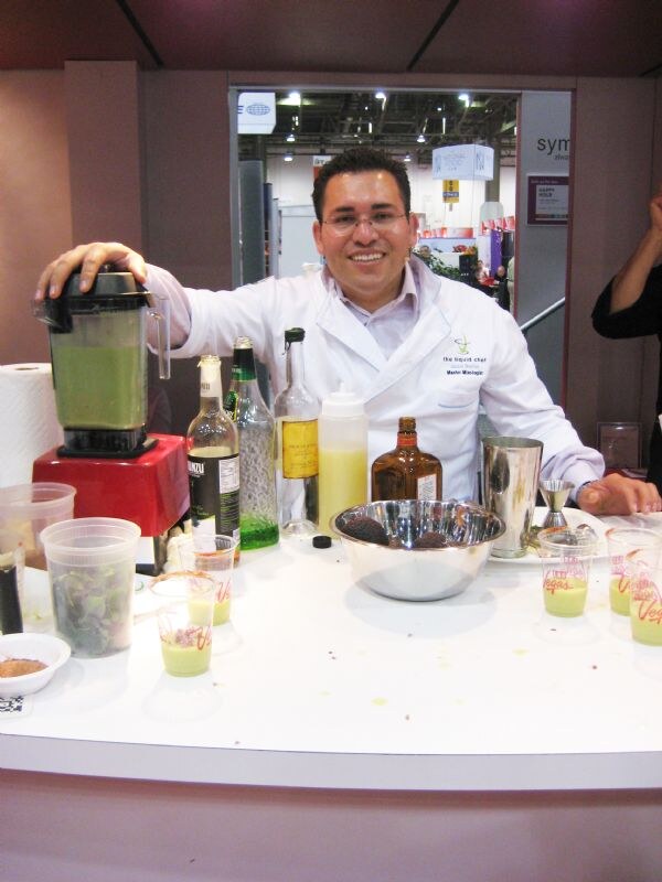 A smiling man in a white chef coat with glasses rests a hand on a blender of green liquid at a counter with bottles, a mixing bowl and small cups of green drink.