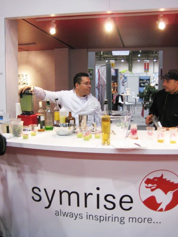 A man in a white chef coat works at a blender on a Symrise counter with bottles and small cups of drinks. Another man in a black cap stands at right.