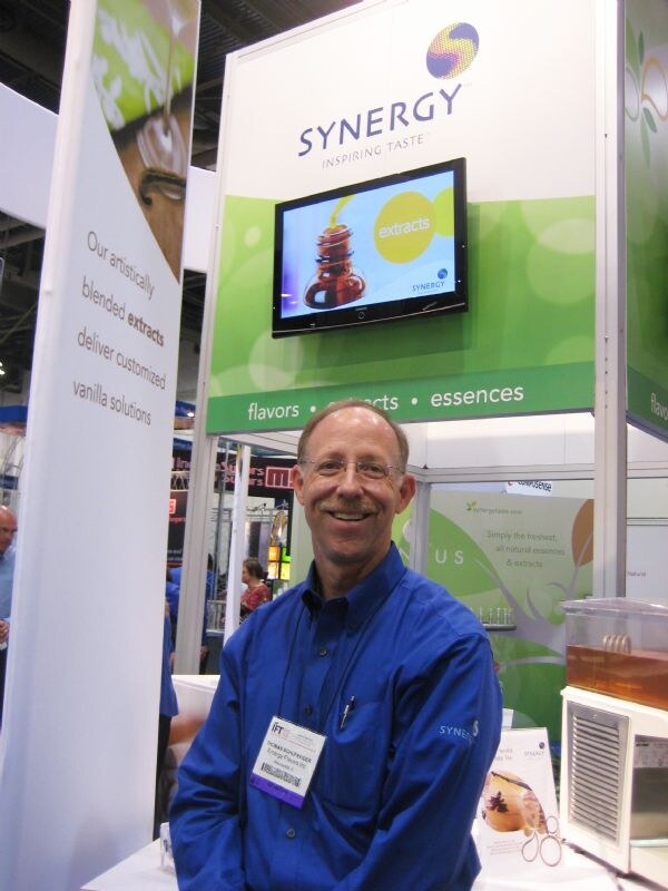 A smiling man with glasses and a blue shirt wears an IFT badge in front of a Synergy booth with a screen reading extracts and a sign for flavors, extracts and essences.
