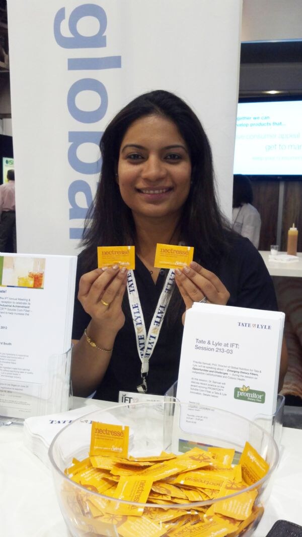 A smiling woman with a Tate & Lyle lanyard holds up two orange Nectresse sweetener packets. A bowl of Nectresse packets and a Tate & Lyle Promitor flyer sit on the table in front of her.