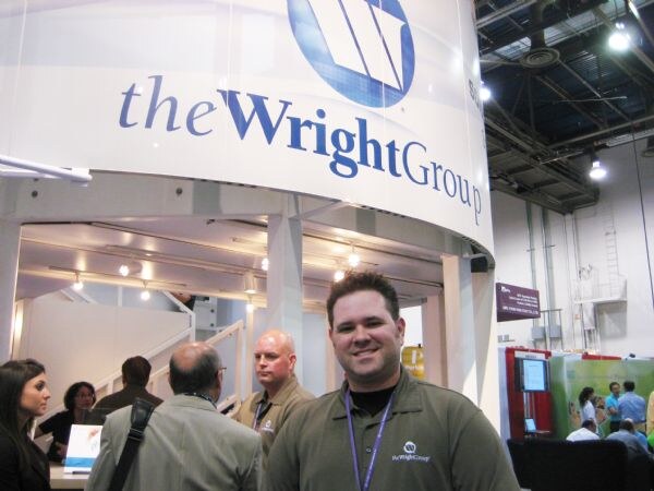 A smiling man in an olive polo shirt with a the Wright Group logo and lanyard stands in front of the company's booth sign. Other attendees stand behind him.