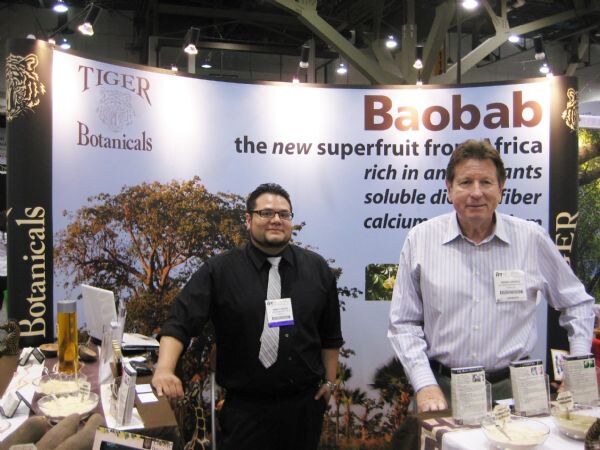 Two men stand at the Tiger Botanicals booth in front of a backdrop reading Baobab, the new superfruit from Africa. Product samples and brochures sit on the tables around them.