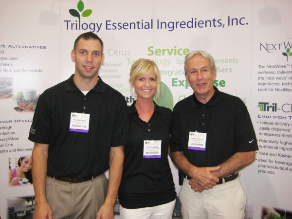 Two men and a woman in black polo shirts with show badges stand together at the Trilogy Essential Ingredients, Inc. booth. The backdrop includes NextWave and Tril-Clear signage.