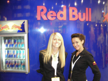 Two women in black stand in front of a blue Red Bull sign and a lit refrigerator cooler stocked with Red Bull cans.