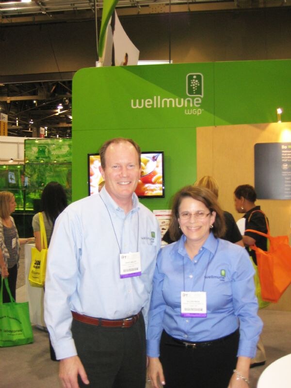A man and a woman in light blue Wellmune shirts with show badges smile in front of the green Wellmune WGP booth. Other attendees carrying tote bags are visible behind them.