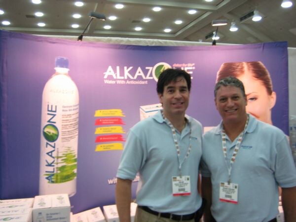 Two men in light blue polo shirts and show badges stand in front of a purple Alkazone banner showing a tall bottle of Alkazone water. Cases of product sit at the bottom of the frame.