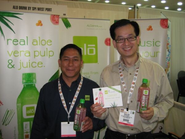 Two smiling men with show lanyards stand at the alō aloe vera drink booth holding bottles. One also holds a card. Banners read real aloe vera pulp and juice.