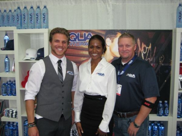 Two men and a woman wearing Aqua Hydrate branded clothing stand together at the booth. Shelves of Aqua Hydrate water bottles and caps are behind them.