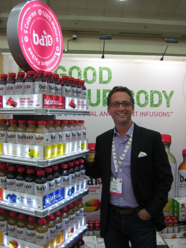A smiling man in glasses and a dark blazer stands beside a tiered, lit Bai display of antioxidant infusion bottles in several flavors, under a round pink Bai sign.
