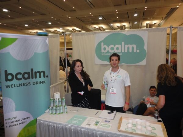 Two people stand behind a table at the bcalm wellness drink booth, with a bcalm banner at left, a cloud-shaped bcalm sign behind them, and cans on the table.