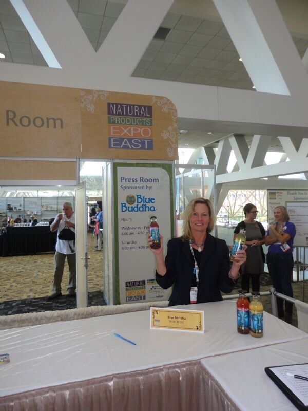 A smiling woman holds up two Blue Buddha bottles at the Press Room entrance, which is sponsored by Blue Buddha. More bottles and a name placard sit on the table.