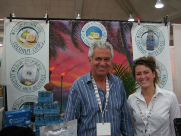 A smiling man and a smiling woman, both wearing lanyards, pose in front of Blue Monkey banners for coconut chips, coconut milk beverage and coconut water.