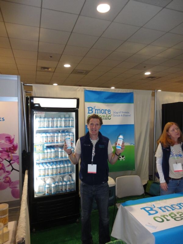 A man in a navy vest holds up two bottles of B'more Organic drink beside a lit refrigerator full of bottles. A woman in a matching vest stands at the right, next to a B'more Organic banner.
