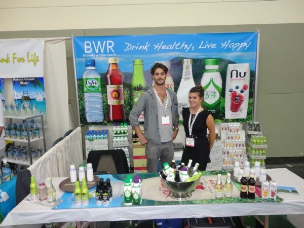 A man and a woman stand behind a table of bottled drinks at the BWR booth. The banner reads 'Drink Healthy, Live Happy' and shows Volvic and Nu Smoothie.