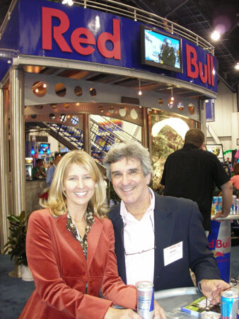 A smiling woman in a red jacket and a man in a dark blazer with a name badge stand together in front of a Red Bull booth, with Red Bull cans on the table in front of them.
