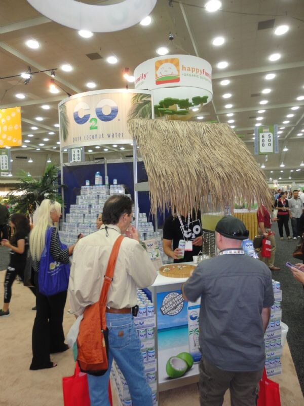 Attendees crowd around the C2O pure coconut water booth, which has stacked cartons and a thatched roof. A Happy Family sign hangs overhead.