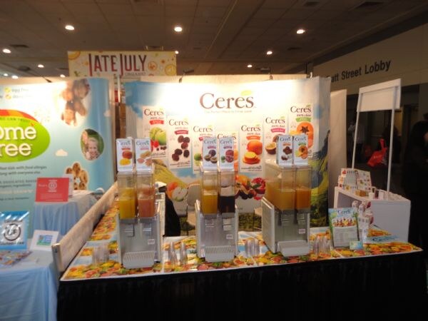 A Ceres booth with a juice-themed backdrop and three juice dispensers filled with orange and dark juices on a fruit-patterned table. No people are visible.