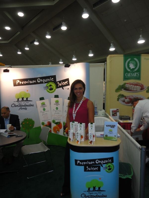 A woman in a pink sleeveless top with a badge stands at the Christodoulou Family premium organic juices booth. A Caesar's booth is visible next door.