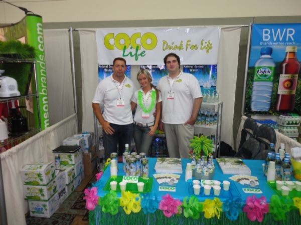 Two men and a woman in white shirts and lanyards stand behind a booth table with green and pink decor, under a Coco Life banner. A BWR banner with Volvic water is on the right.