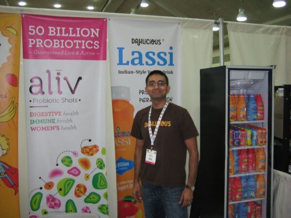 A man in a brown Dahlicious T-shirt and lanyard stands between an aliv Probiotic Shots banner and a Dahlicious Lassi banner, beside a glass-door refrigerator of bottled drinks.