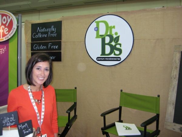 A smiling woman in an orange top and lanyard stands at a booth with a burlap backdrop and a round Dr. B's premium microbrewed tea sign, with two green director's chairs nearby.