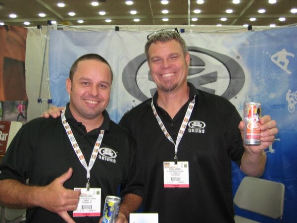 Two smiling men in black EX Drinks shirts and lanyards pose together in front of a blue EX Drinks banner, each holding a can of the drink.